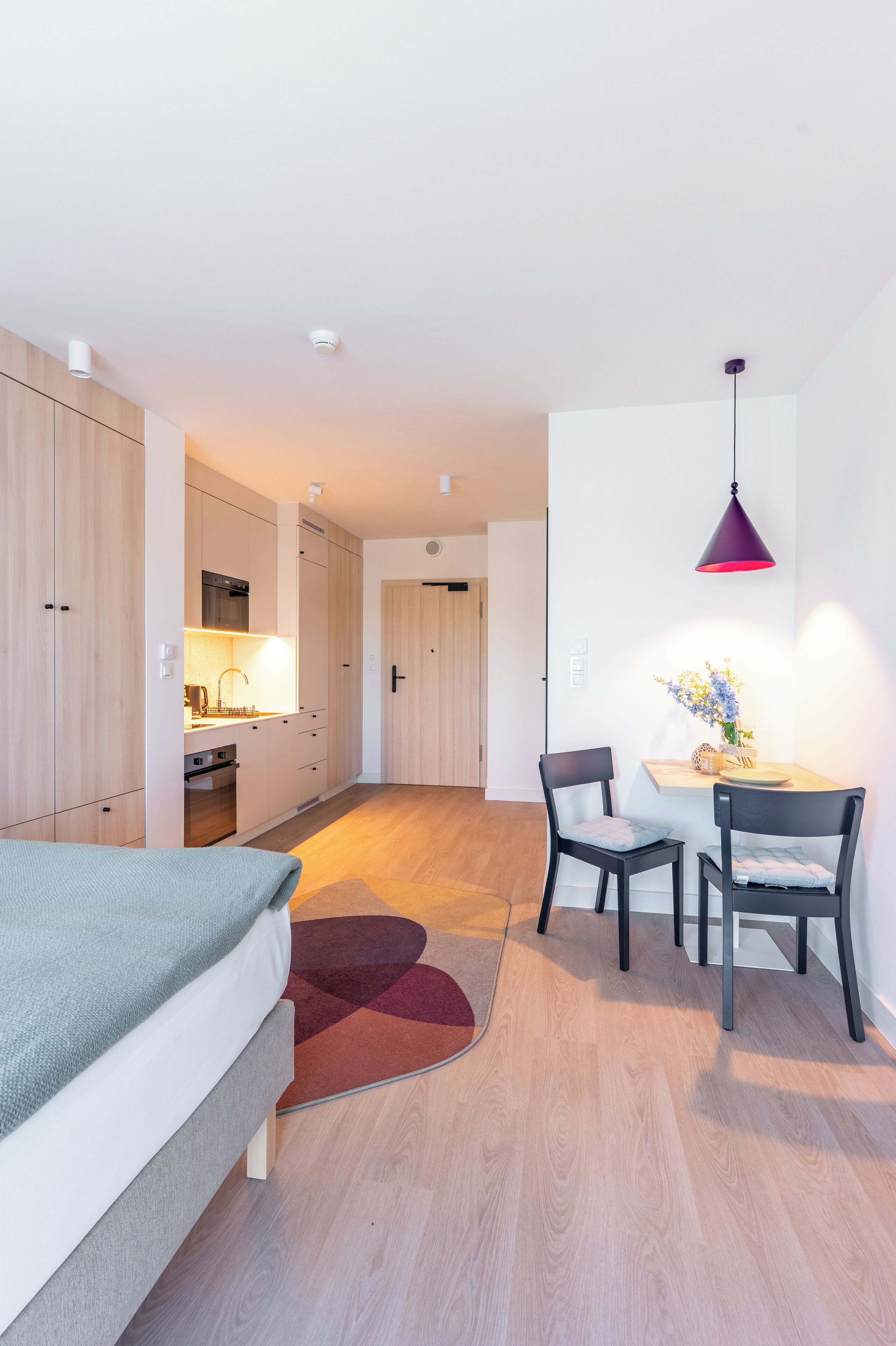 A modern hotel room with a bed, a desk, a chair, a small table with a vase, and a kitchen area.