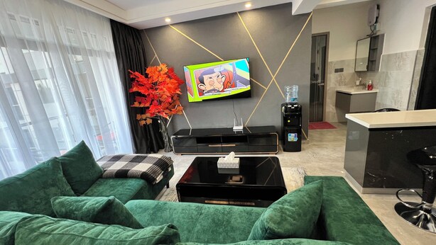 Una sala de estar moderna con un sofá verde, un televisor de pantalla plana, un dispensador de agua y una pequeña cocina.