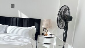 Una cama bien hecha con sábanas blancas, una mesita de noche con lámpara, un ventilador y una alfombra mullida con zapatillas.