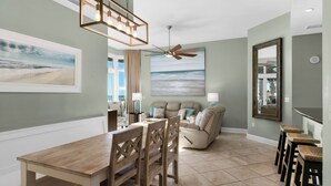 A dining area with a wooden table and chairs, a ceiling fan, and a painting of a beach scene. - Rare First-Floor Gulf-Front Corner Unit | Private Terrace + Beach chairs (Panama City Beach)