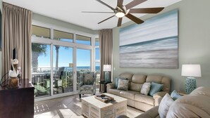 A spacious living room with a large sliding glass door leading to a balcony, a ceiling fan, and a painting of a beach scene. - Rare First-Floor Gulf-Front Corner Unit | Private Terrace + Beach chairs (Panama City Beach)