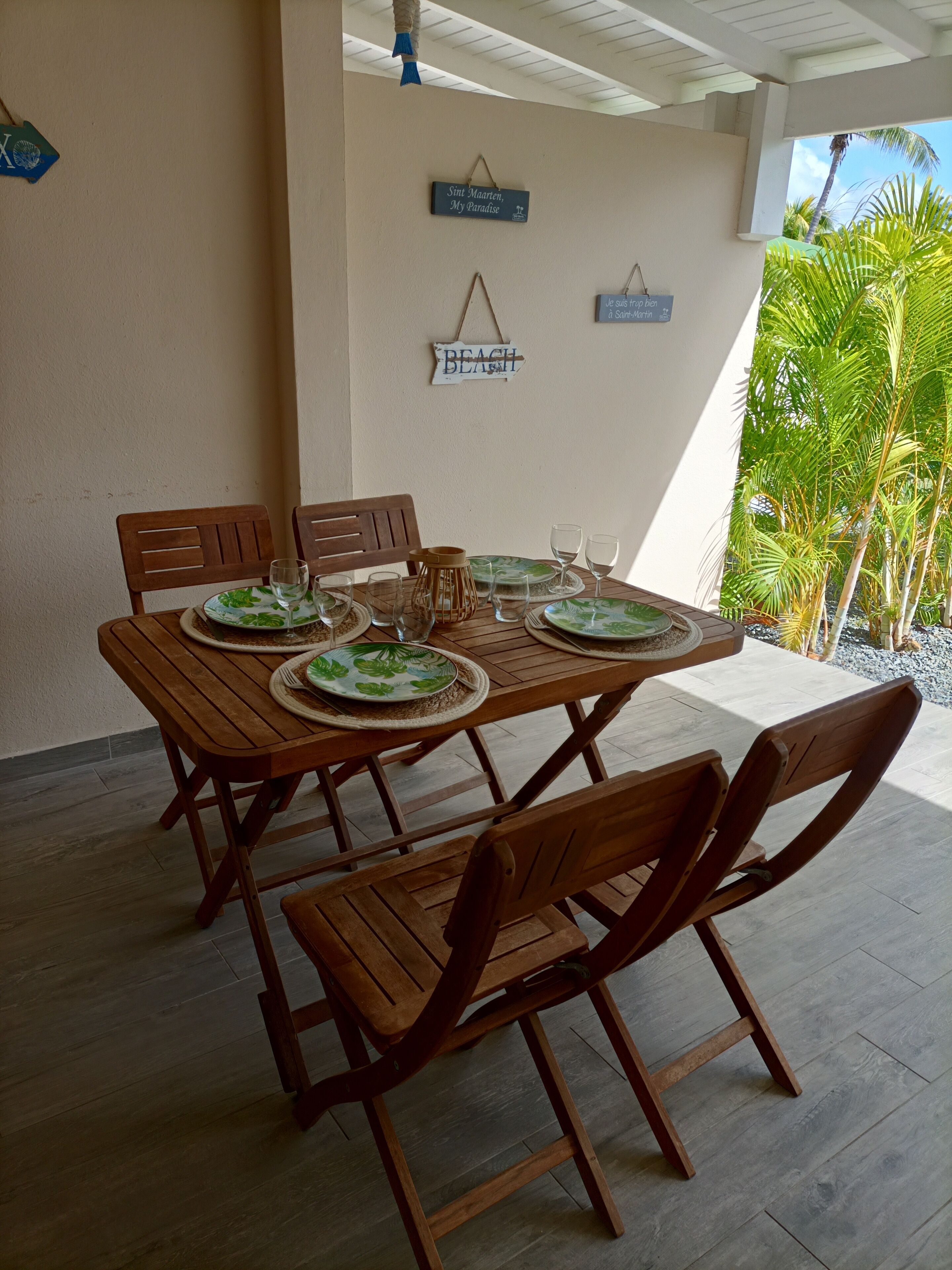 A wooden table set with plates, glasses, and a candle, surrounded by chairs, under a ceiling with hanging decorations, with palm trees visible outside.