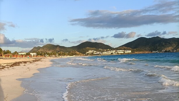 Une plage avec des vagues, un ciel dégagé et des montagnes à l’horizon.