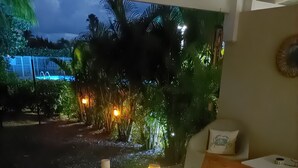 Une table en bois, dressée avec des boissons, des amuse-gueules et une bougie, dans une pièce donnant sur un jardin illuminé la nuit.