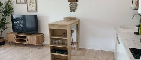 Un salon moderne comprenant un meuble de rangement en bois pour l’équipement audiovisuel, un téléviseur à écran plat, une petite table en bois avec un panier, et un coin cuisine doté d’un plan de travail et d’un évier blancs.