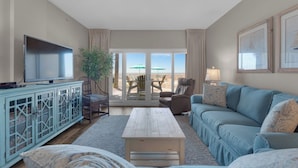 A living room with a television, a blue sofa, a wooden coffee table, a cabinet with glass doors, a plant, and a view of the beach through large sliding doors. - Sandy Key 123:  2 BR / 2 BA Gulf Front Condo in Perdido Key, FL  Sleeps 6 (Pensacola)