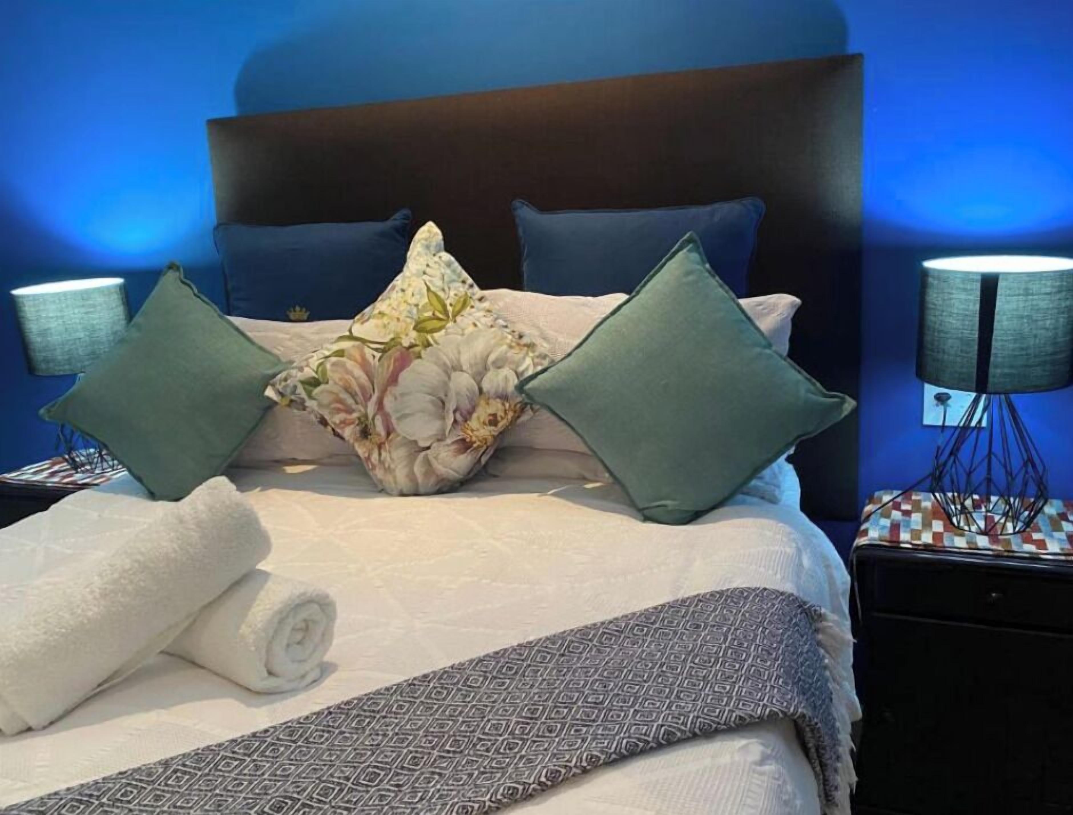 A neatly made bed with floral and solid pillows, a rolled white towel, and a patterned throw. There are two bedside tables with lamps and a small decorative item on one.