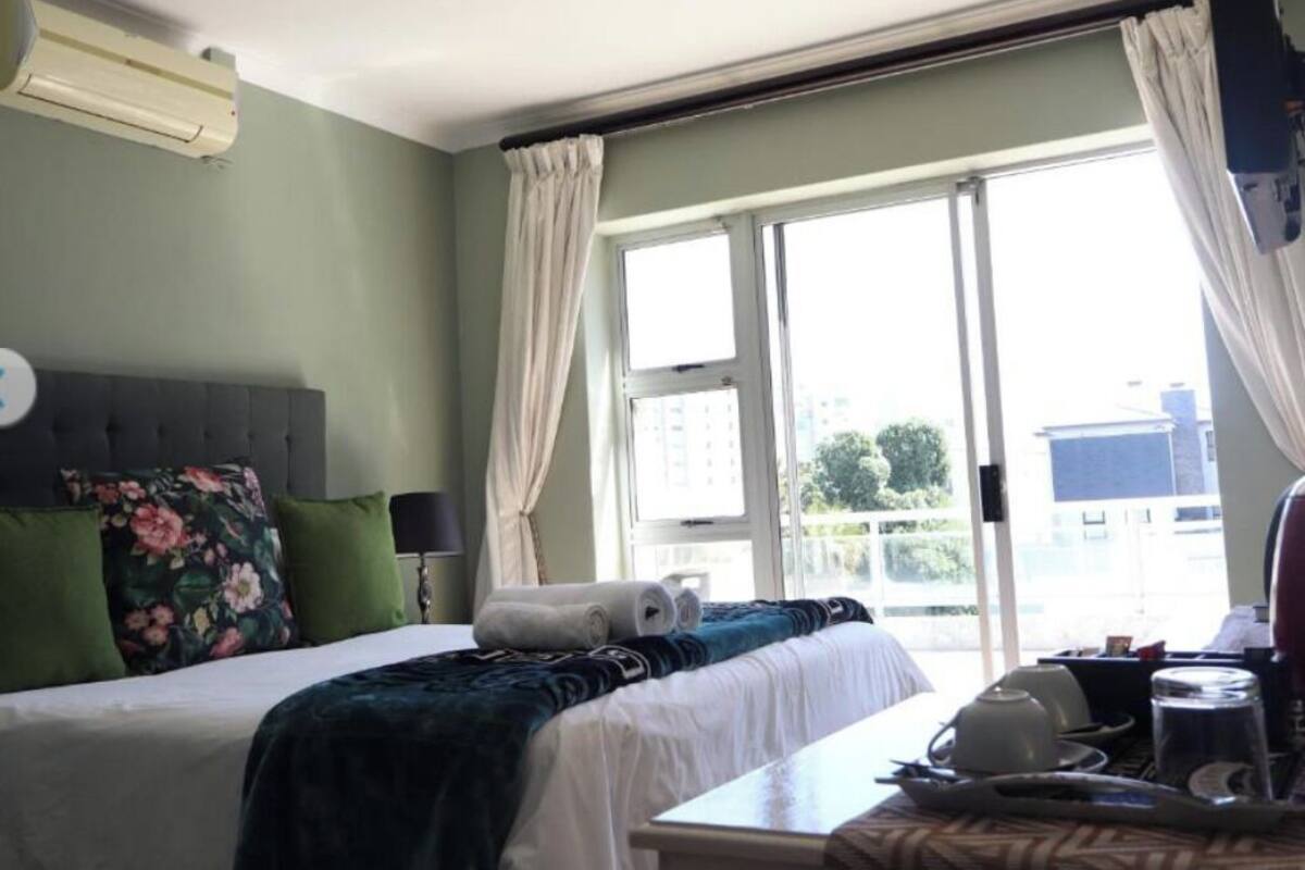 A hotel room with a bed, a table, a window with curtains, and an air conditioning unit.