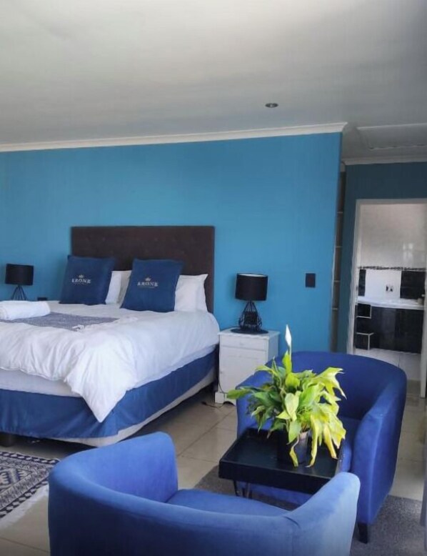 A hotel room with a large bed, two bedside tables with lamps, a blue armchair, a small table with a plant, and a kitchenette in the background.