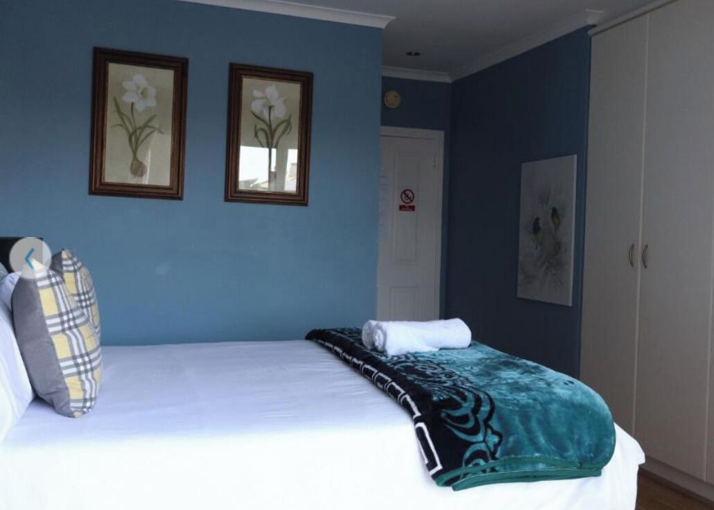 A neatly made bed with a white comforter and a teal blanket, two framed floral paintings on the wall, a closed door with a 'No Smoking' sign, and a wardrobe to the right.