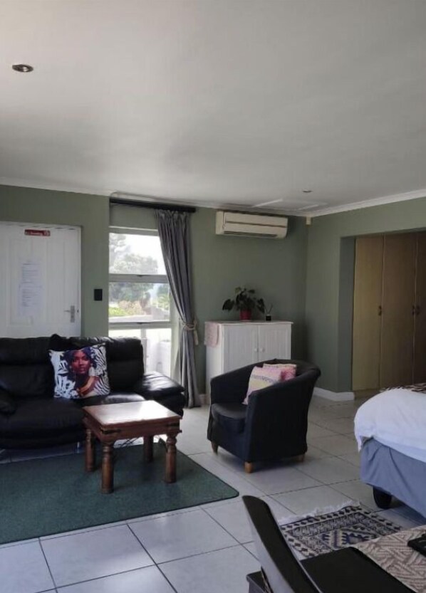 A room with a black leather sofa, a wooden coffee table, a dark armchair, a bed with white bedding, a white cabinet, and an air conditioning unit.