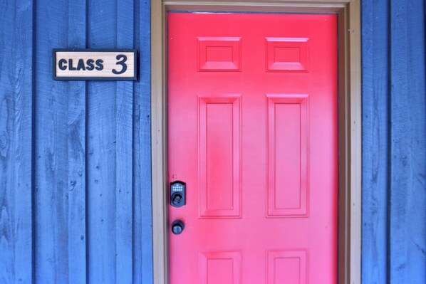 A bright pink door with a "CLASS 3" sign above it, set against a blue wooden background.