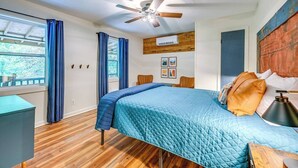 A bedroom with a bed, a ceiling fan, a window with curtains, a wooden floor, and a wall-mounted air conditioner.