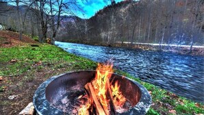 A campfire in a metal bowl with a river and forest in the background.