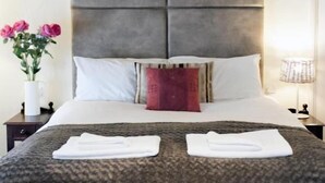 A modern hotel room with a large bed, a grey headboard, white bedding, a red accent pillow, a vase with pink flowers, and a lamp on the nightstand. - Woolpack Inn (Holmrook)