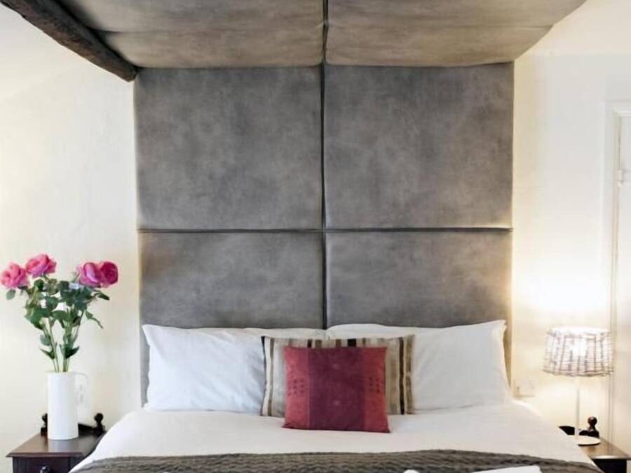 A modern hotel room with a large bed, a grey headboard, white bedding, a red accent pillow, a vase with pink flowers, and a lamp on the nightstand.