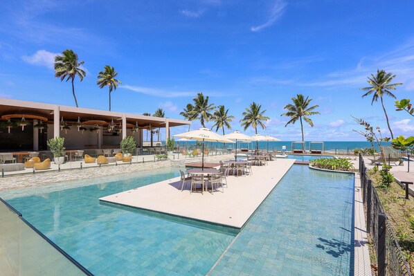 A pool area with lounge chairs and umbrellas, surrounded by palm trees and a view of the ocean. - Cais Ecoresidencia Luxo em Muro Alto (Ipojuca)