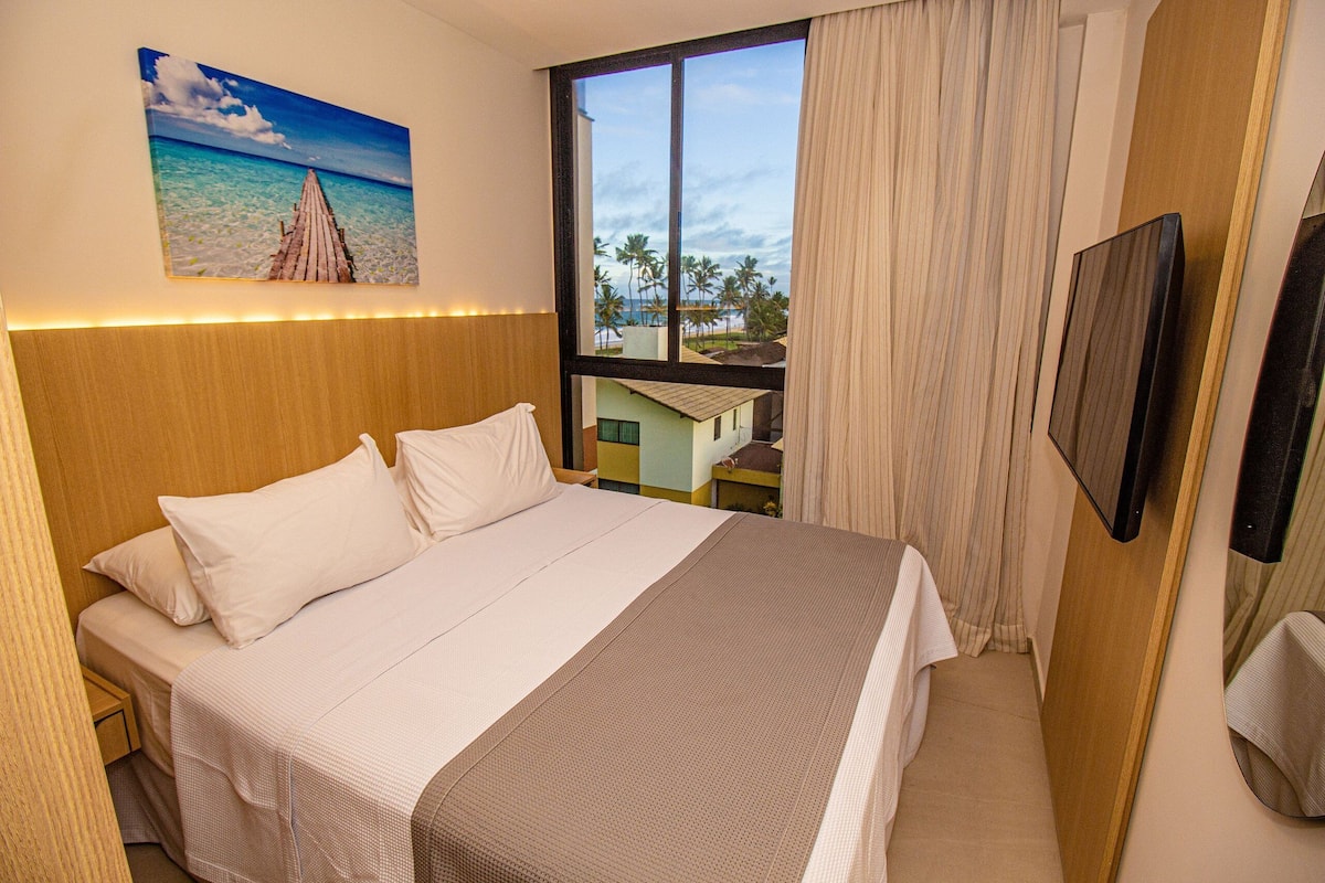 A hotel room with a bed, a painting of a beach, a window with curtains, and a television.