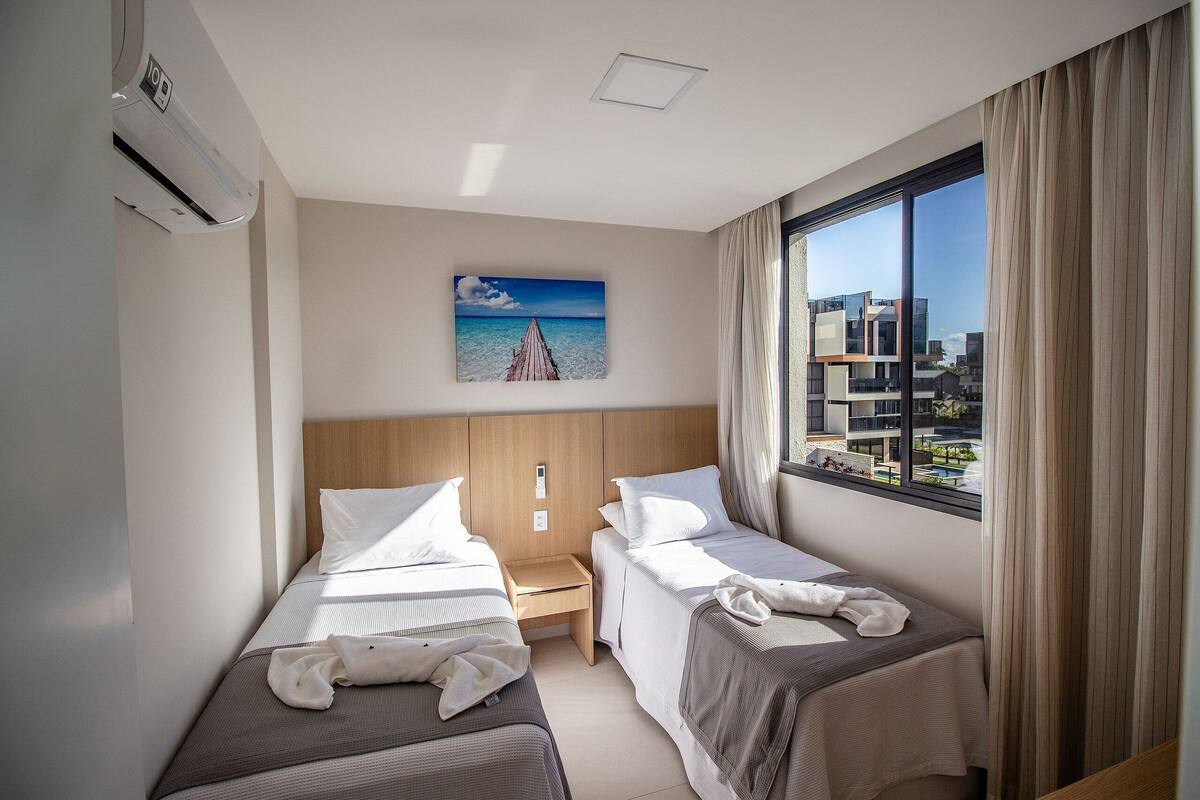 A hotel room with two beds, a nightstand, a painting of a beach, and a view of buildings through the window.