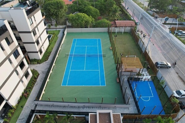 An aerial view of a tennis court surrounded by buildings, with a street and vehicles nearby. - Cais Ecoresidencia Luxo em Muro Alto (Ipojuca)