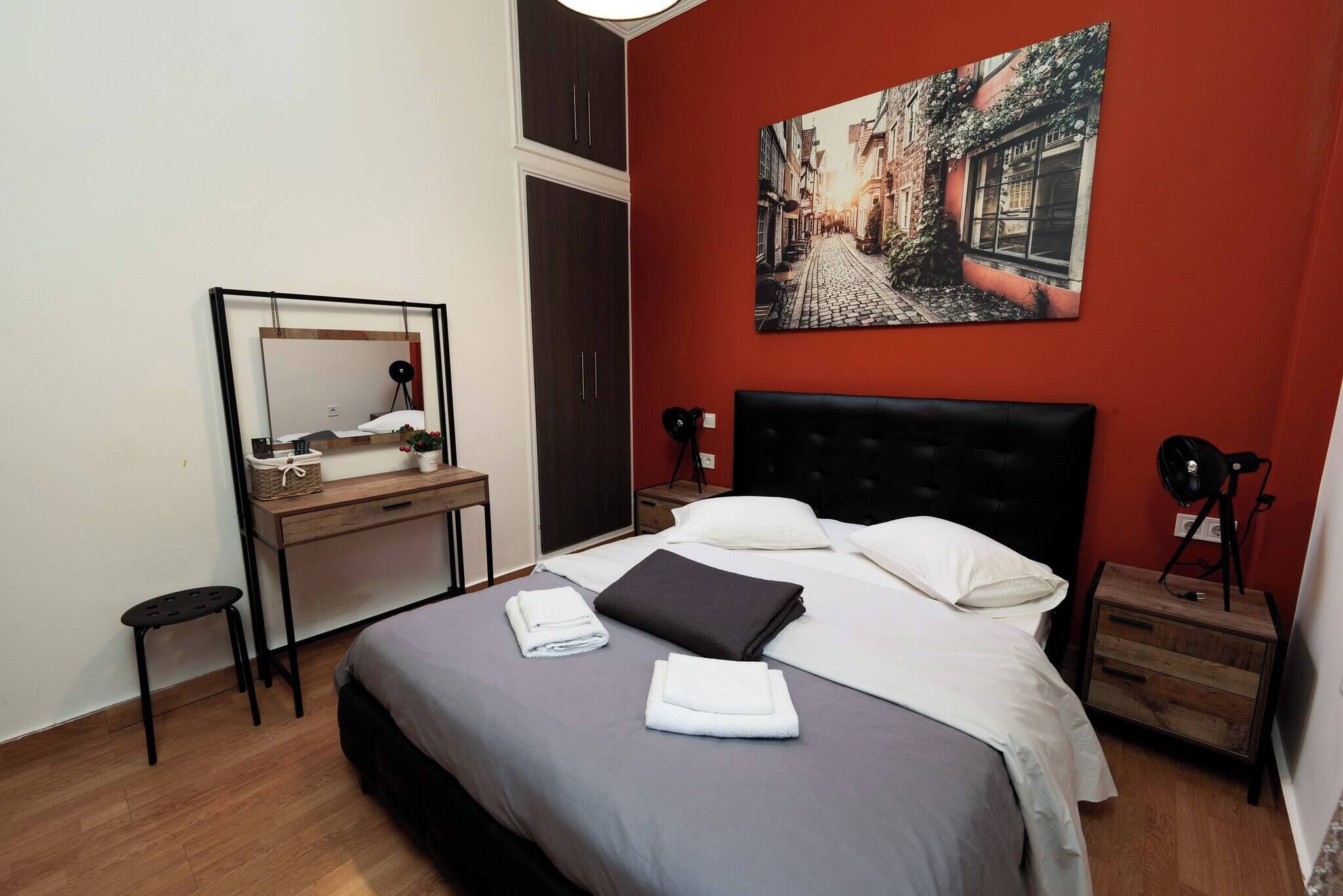A neatly made bed with pillows and folded towels, a wooden desk with a mirror, and a red wall with a street scene painting.