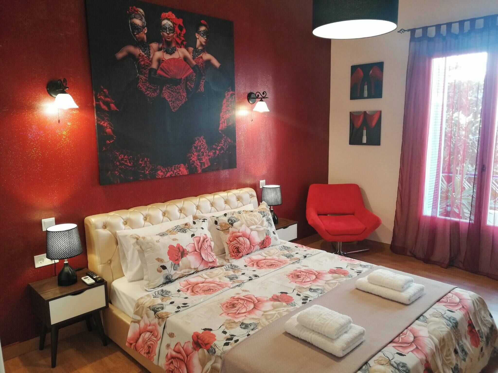 A hotel room with a tufted headboard, floral bedspread, two bedside lamps, a red armchair, and a large painting on the wall.