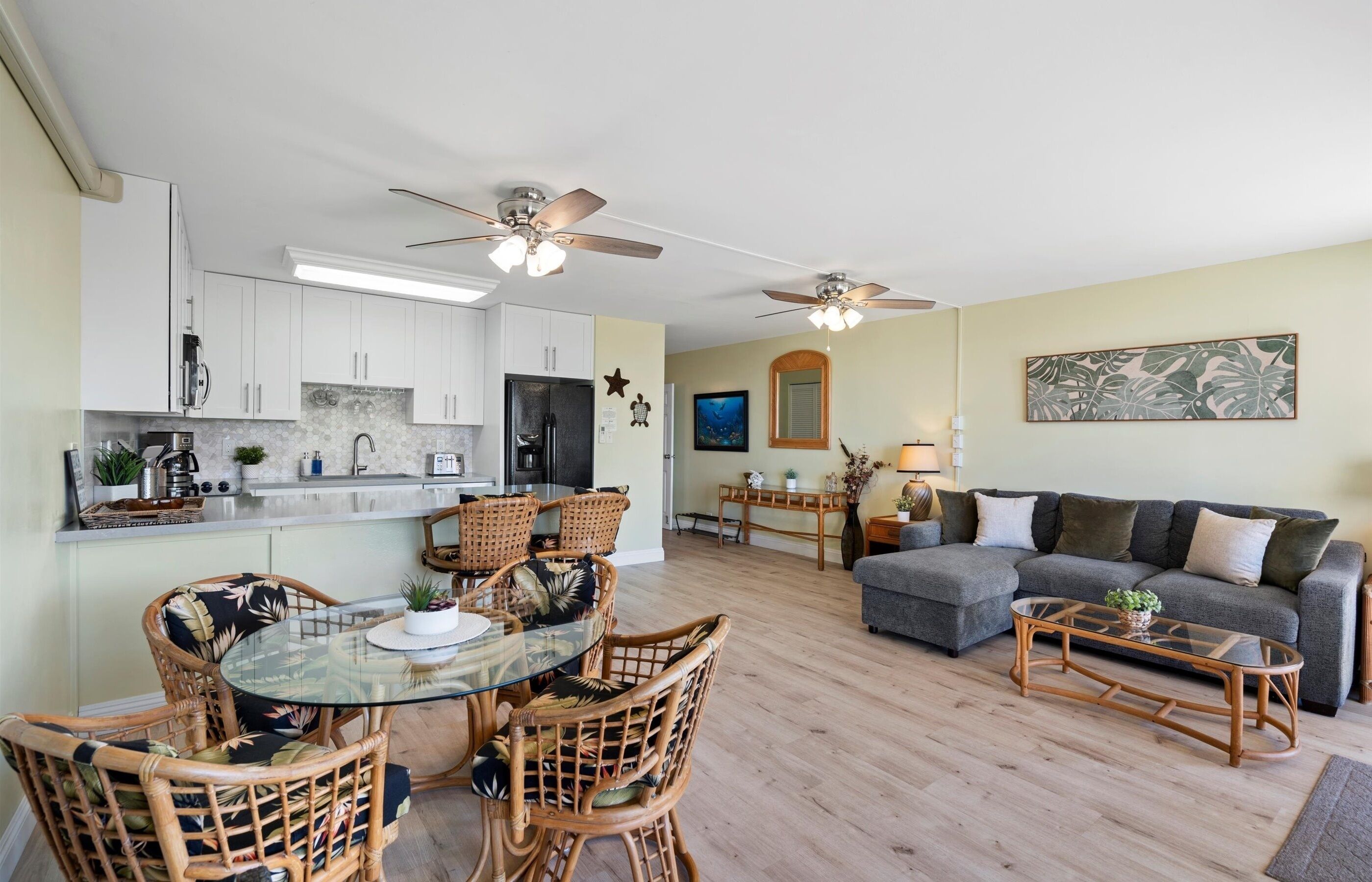 A modern kitchen with white cabinets, a dining area with wicker chairs, a gray sofa, and a ceiling fan.