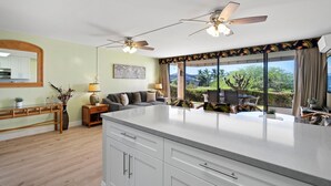 A spacious living area with a large island, ceiling fans, a dining table, and a view of the outdoors. - Kihei Ali'i Kai D108 2 Bedroom Condo (Kihei)
