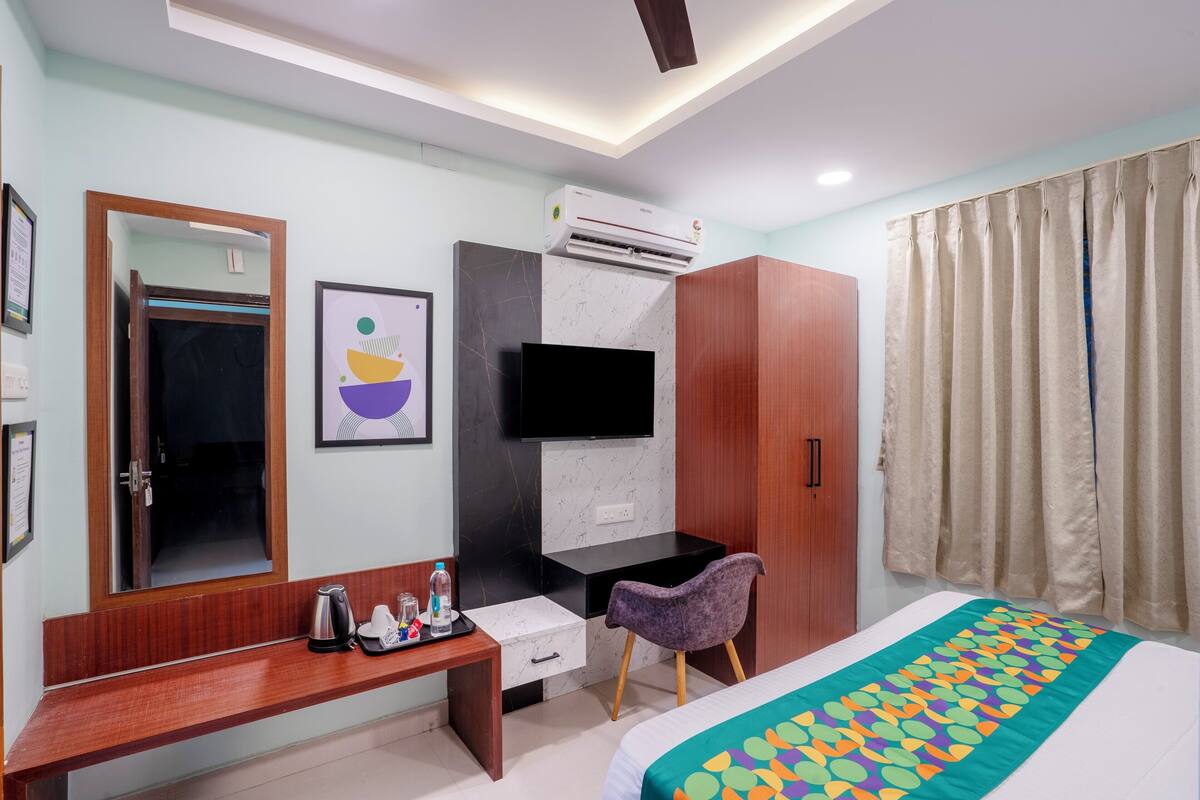 A hotel room with a wooden bench, a desk with a flat-screen TV, a wardrobe, a bed with a colorful bedspread, and a window with curtains.