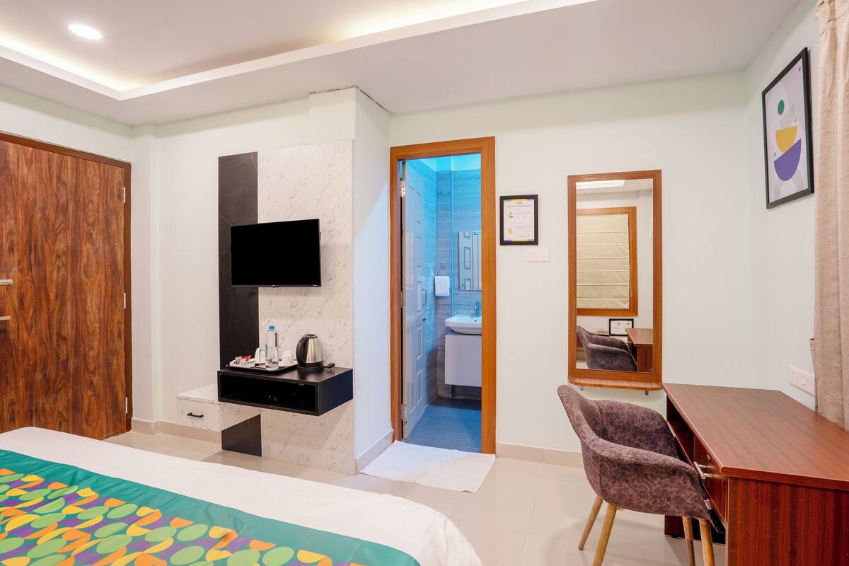 A hotel room with a bed, a desk, a chair, a TV, a mirror, and a bathroom visible through an open door.
