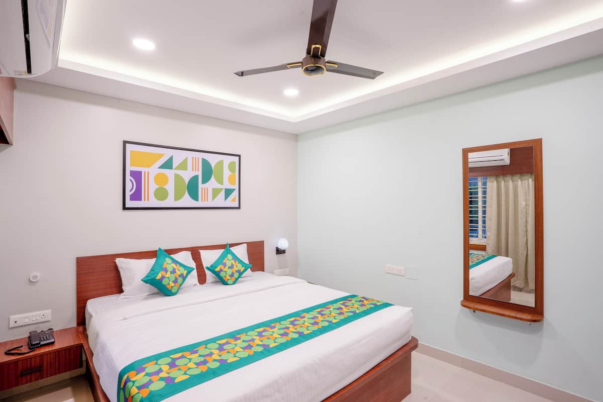 A hotel room with a bed, bedside table, nightstand, wall-mounted air conditioner, ceiling fan, and a framed abstract artwork.