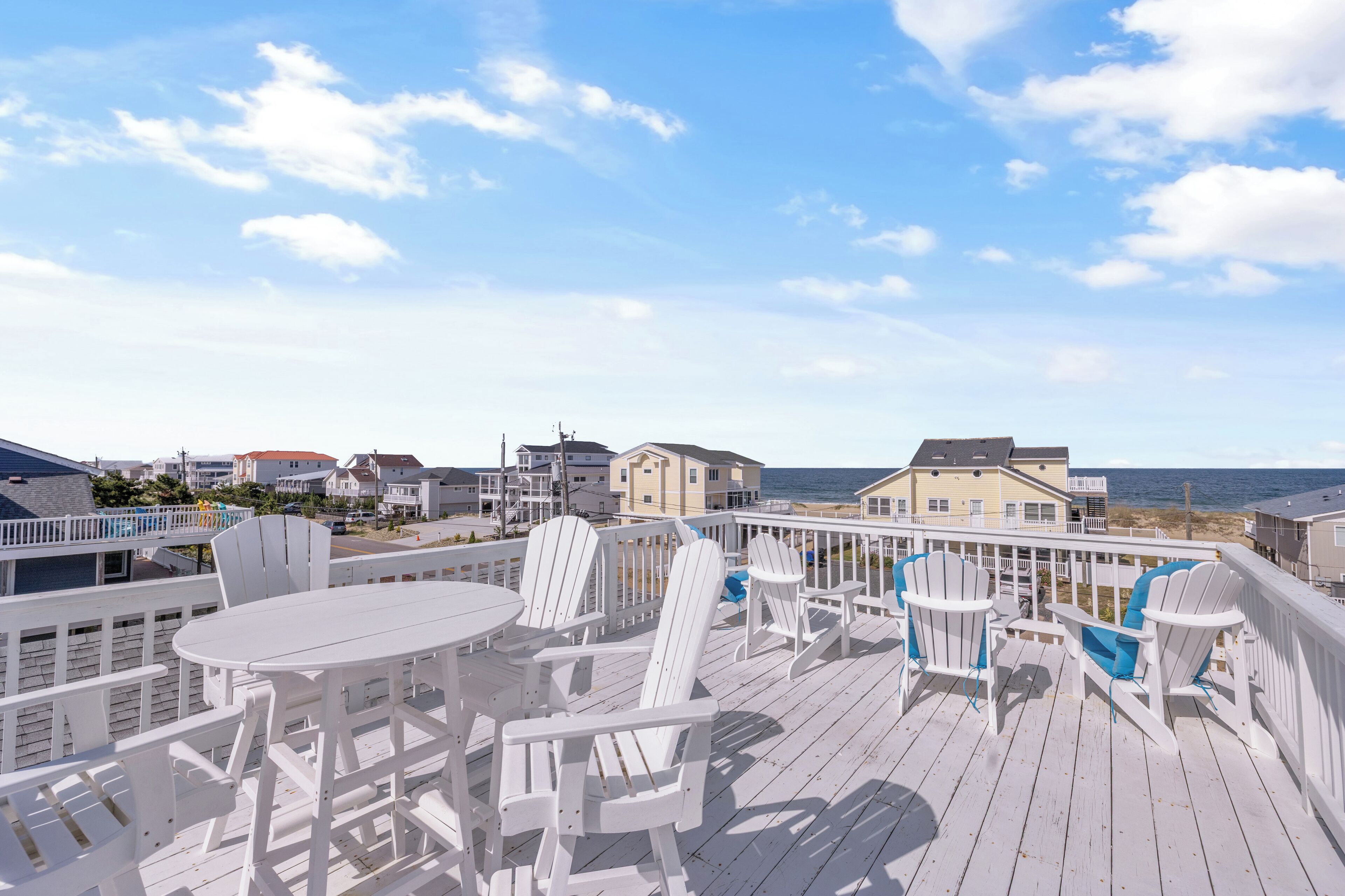 A wooden deck with white chairs and tables, overlooking a beach and buildings.