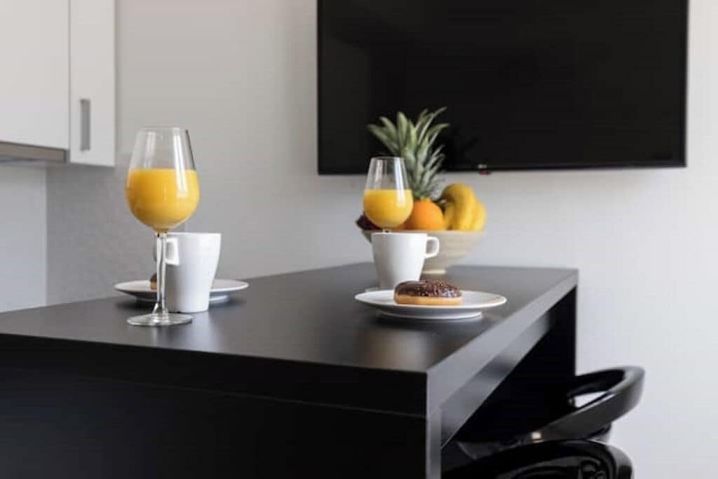 A modern kitchen with a black countertop, a white mug, a glass of orange juice, a plate with a donut, and a bowl of fruit including a pineapple and bananas.