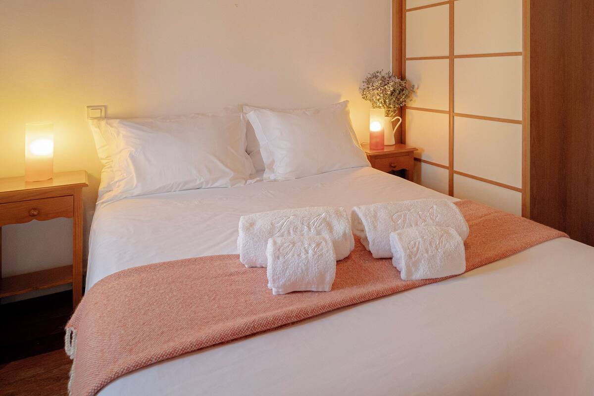 A neatly made bed with white linens, a pink throw blanket, and two folded white towels. There is a bedside table with a lamp and a vase of flowers.