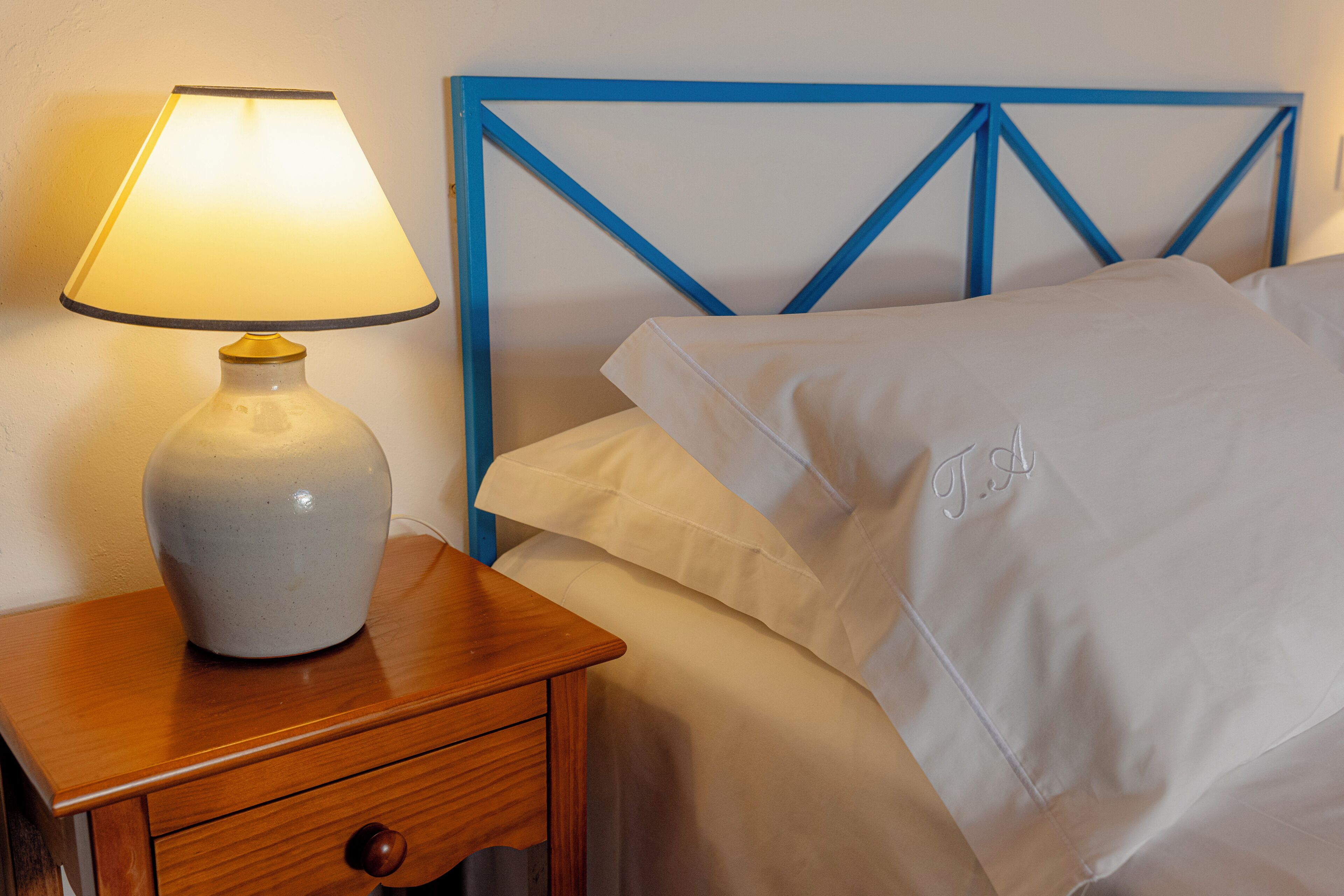 Una cama con cabecera azul, una lámpara de noche y un almohadón blanco con iniciales bordadas.