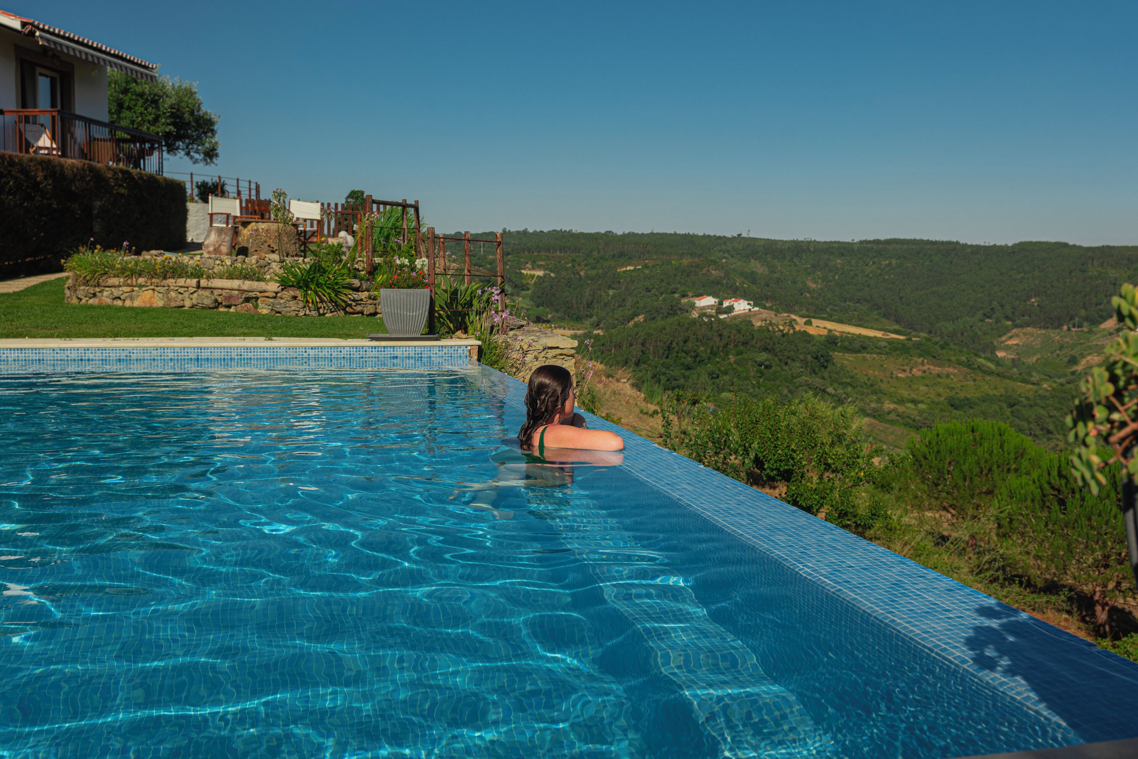 A swimming pool with clear blue water, surrounded by greenery and a view of a hilly landscape.