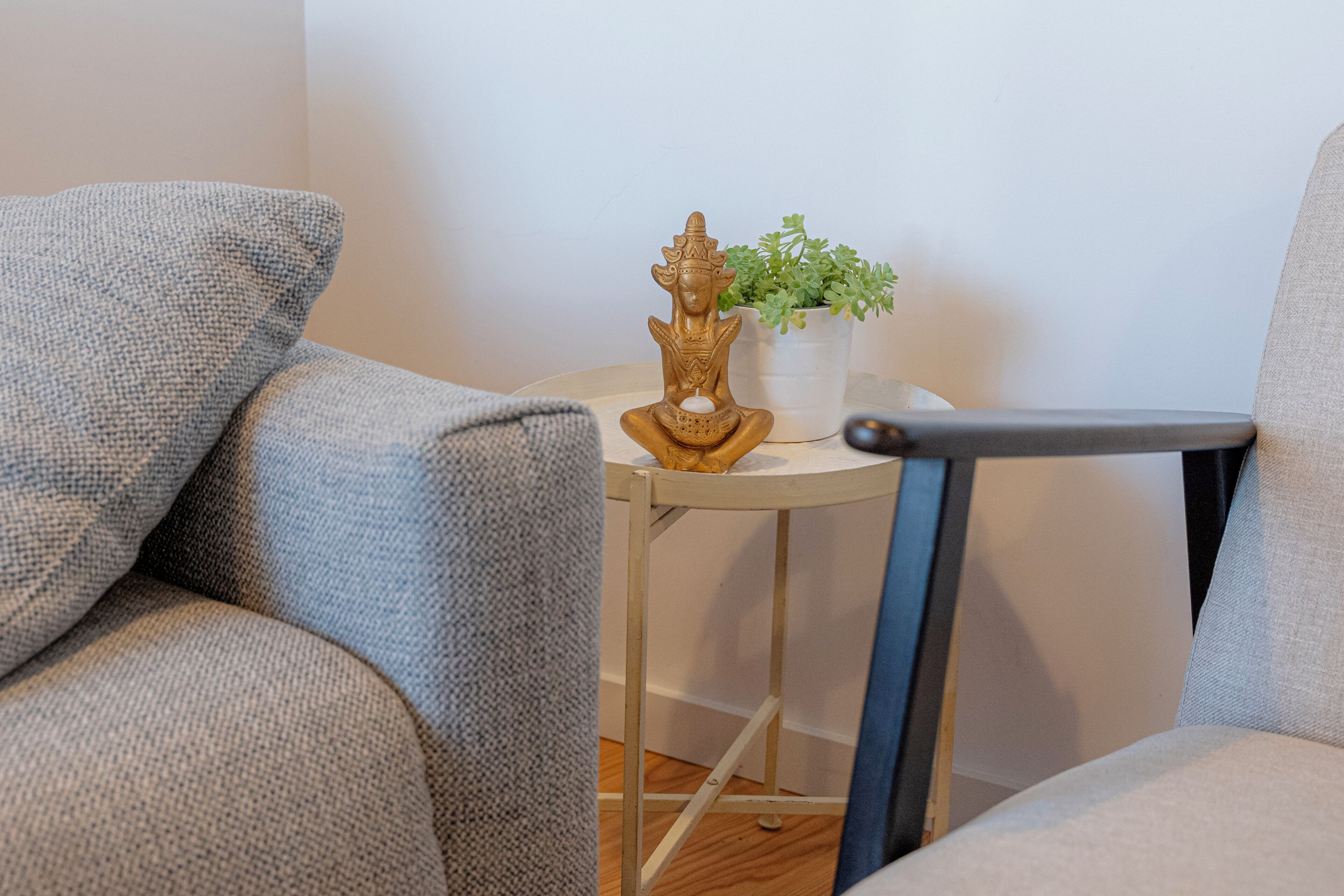 Et hjørne af et rum med en grå sofa, et rundt bord med en plante og en statue, samt en stol med en sort armlæn.
