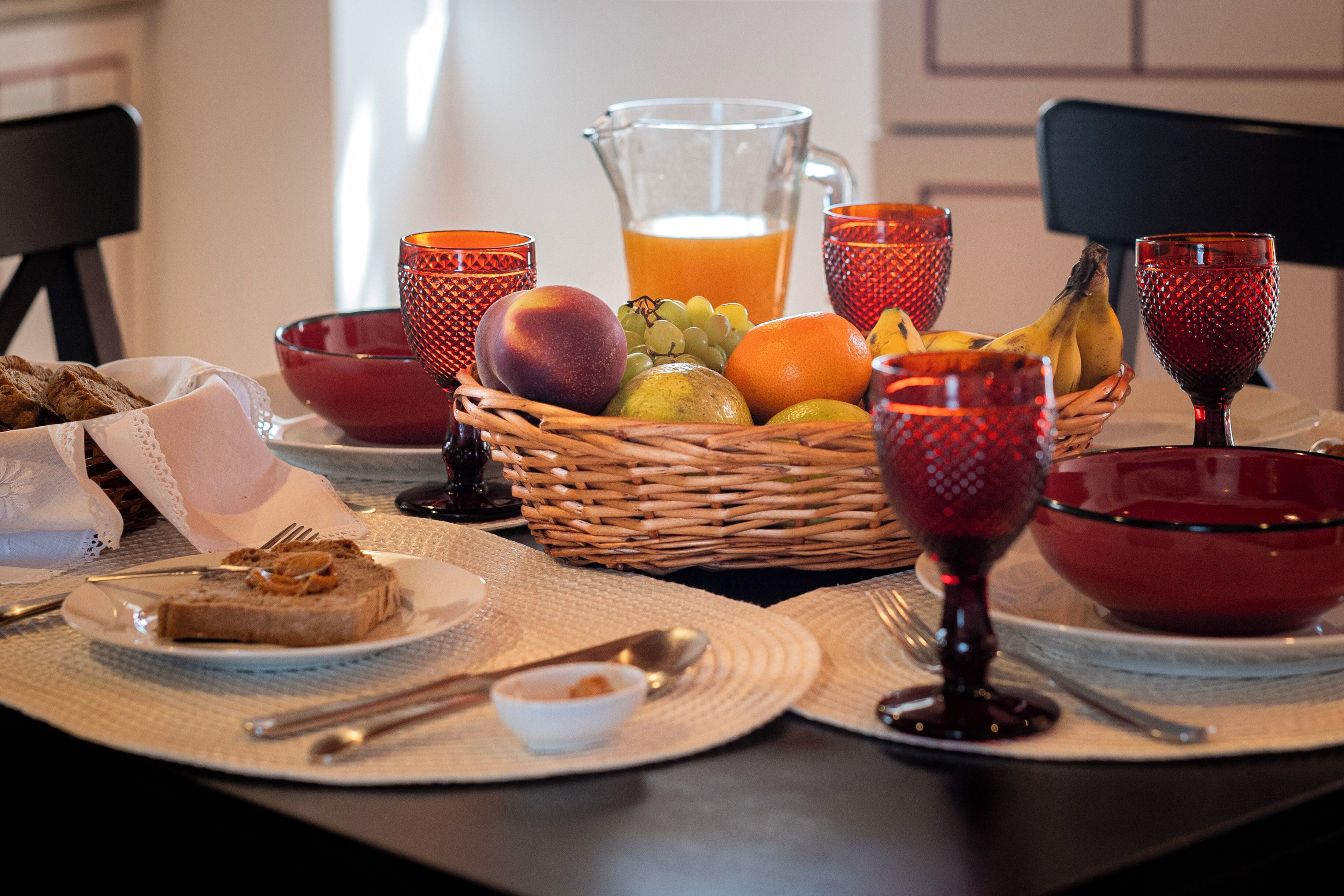 Een ontbijttafel met een fruitmand, een karaf sinaasappelsap en rode glazen.