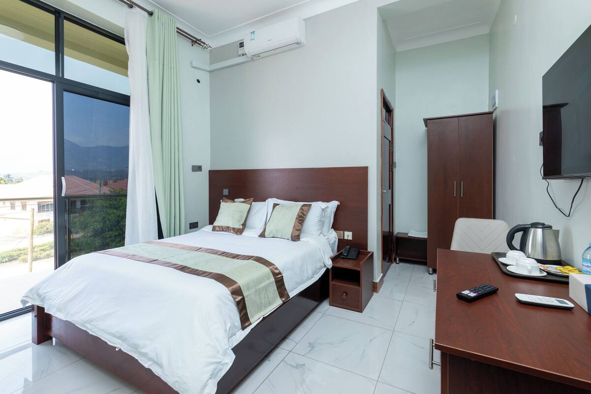 A hotel room with a bed, a desk with a coffee maker, a TV, and a window with curtains.