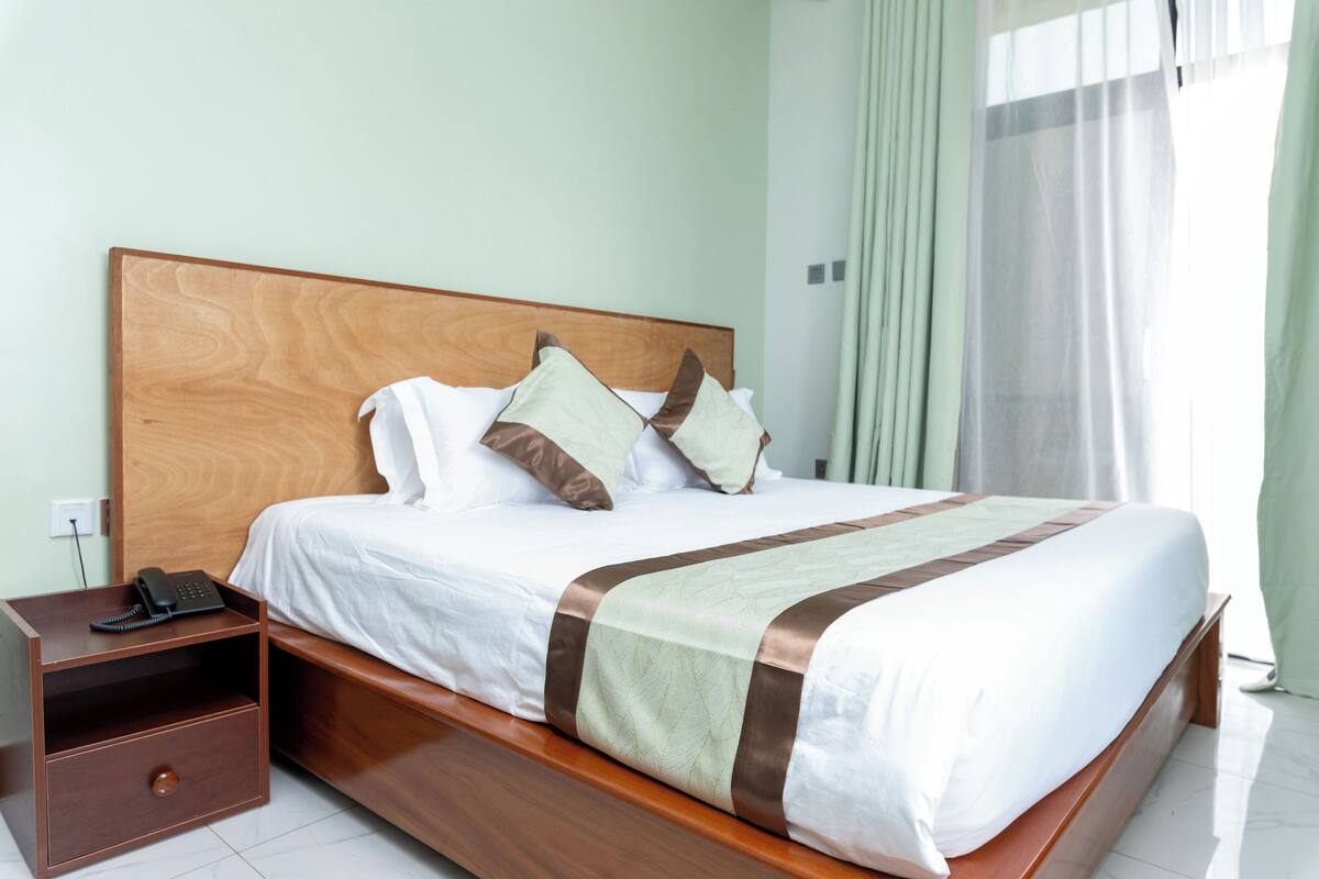 A neatly made bed with a wooden headboard, a bedside table with a telephone, and a window with curtains.