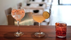 Een bar met drie drankjes op een houten tafel, een rieten lamp erboven en een wazige achtergrond die een eetgedeelte doet vermoeden.