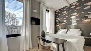 Een slaapkamer met een bed, een bureau, een stoel en uitzicht op gebouwen door een raam.