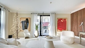 Een moderne woonkamer met een witte bank, een ronde salontafel en een witte fauteuil. Er is een groot raam met gordijnen, een potplant en een vaas.