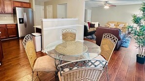 Un coin repas avec une table en verre entourée de chaises en osier, un réfrigérateur en acier inoxydable, des meubles de rangement en bois et un ventilateur de plafond en arrière-plan.