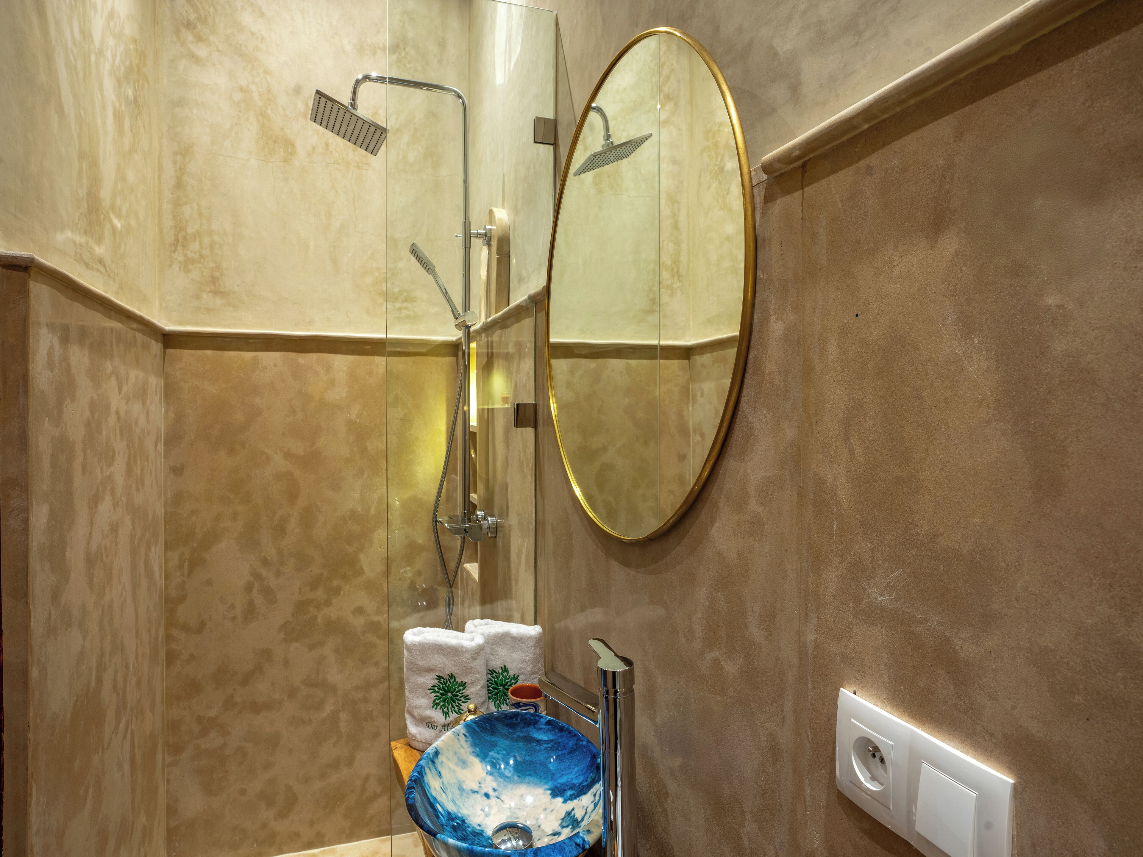 A bathroom with a glass-enclosed shower, a round mirror, a sink with a blue basin, and two towels on the counter.