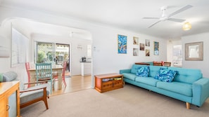 A living room with a teal sofa, wooden coffee table, and a dining area with a table and chairs. - Mirrabooka - Charming 2-bedroom holiday house (Lake Cathie)
