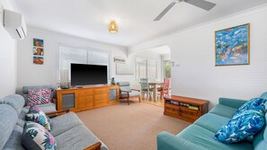 A living room with a flat-screen TV, wooden cabinet, and a dining area with chairs and a table. - Mirrabooka - Charming 2-bedroom holiday house (Lake Cathie)