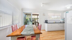 A dining area with a wooden table and chairs, a kitchen with white cabinets, and a view through a glass door to an outdoor area. - Mirrabooka - Charming 2-bedroom holiday house (Lake Cathie)