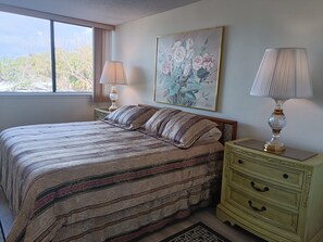 A bedroom with a large bed, a nightstand, a dresser, a window with curtains, and a floral painting on the wall. - Our House at the Beach W-102 (Siesta Key)