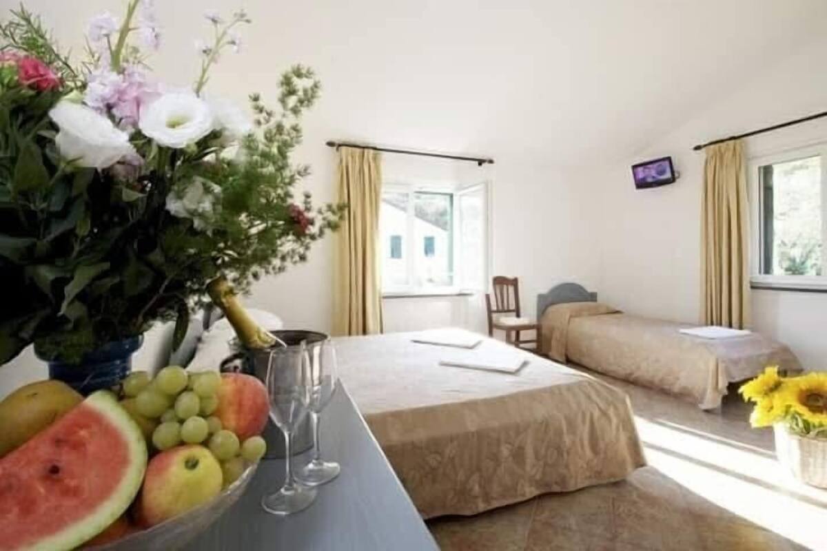Ein Hotelzimmer mit Bett, Nachttisch mit Blumenvase, Obstschale und einem Fenster mit Vorhängen.